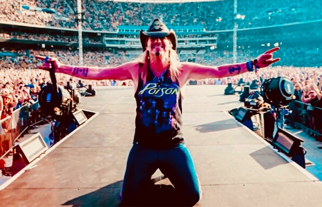 Rock performer on a crowded stadium stage, arms wide open, wearing a hat and a Poison shirt, engaging with the audience far in the stands.