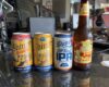 Four beverages on a desk: two yellow Shiner Bock cans, a Shiner IPA can, and a Shiner Seasonal Raspberry Lemonade bottle in front of office equipment.