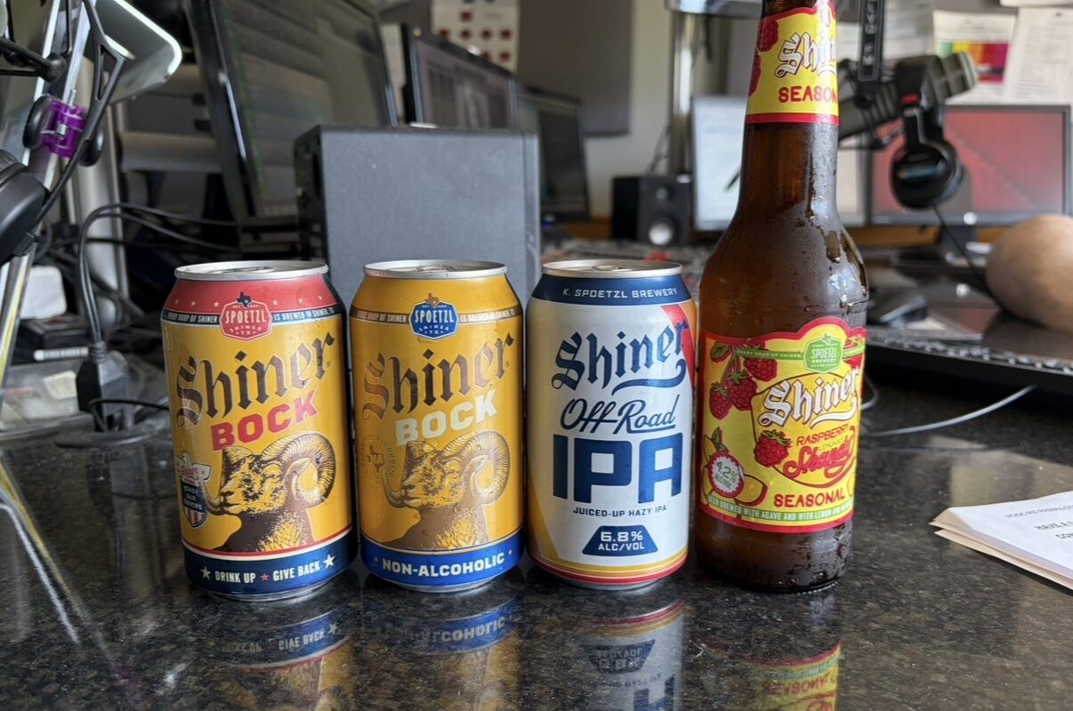 Four beverages on a desk: two yellow Shiner Bock cans, a Shiner IPA can, and a Shiner Seasonal Raspberry Lemonade bottle in front of office equipment.